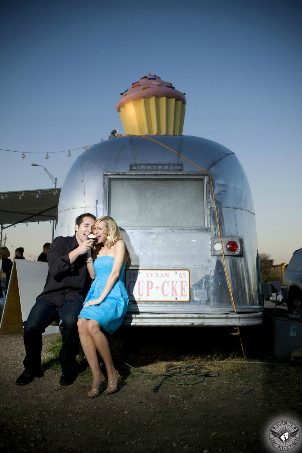 hot blond girl with blue dress and tan heels shares a cupcake with a dark haired guy wearing a dark blue shirt and black pants sitting together on the bumper of the original hey cupcake silver airstream travel trailer with a large cupcake on top in this engagement photograph in southern austin  